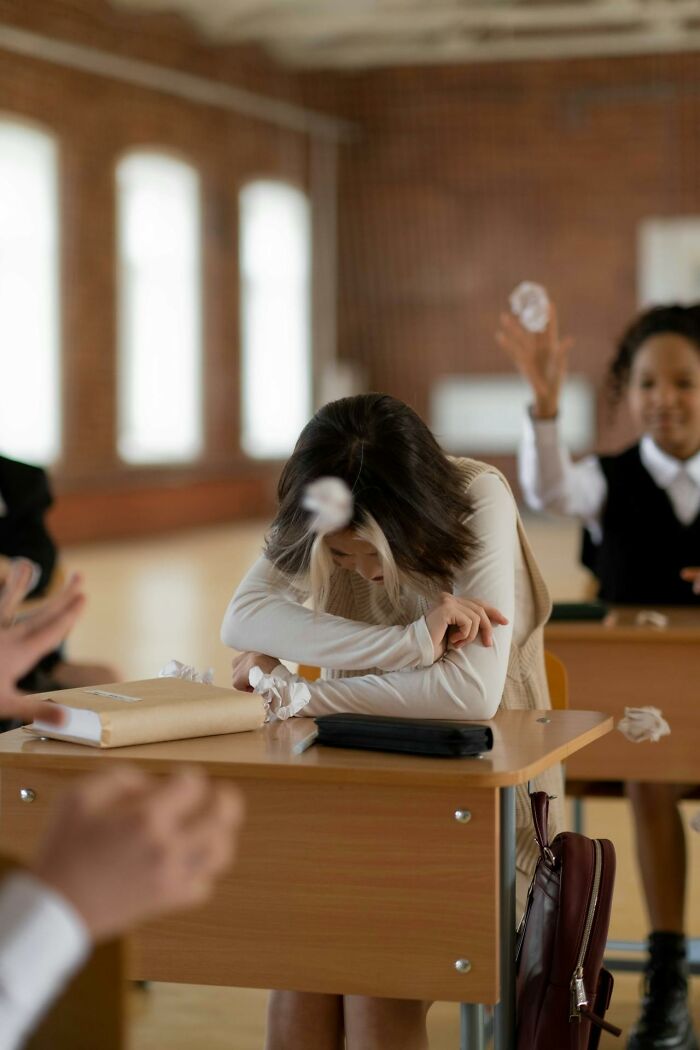 Student covering head at desk while classmates throw paper balls in classroom illustrating toxic friendships. - 43