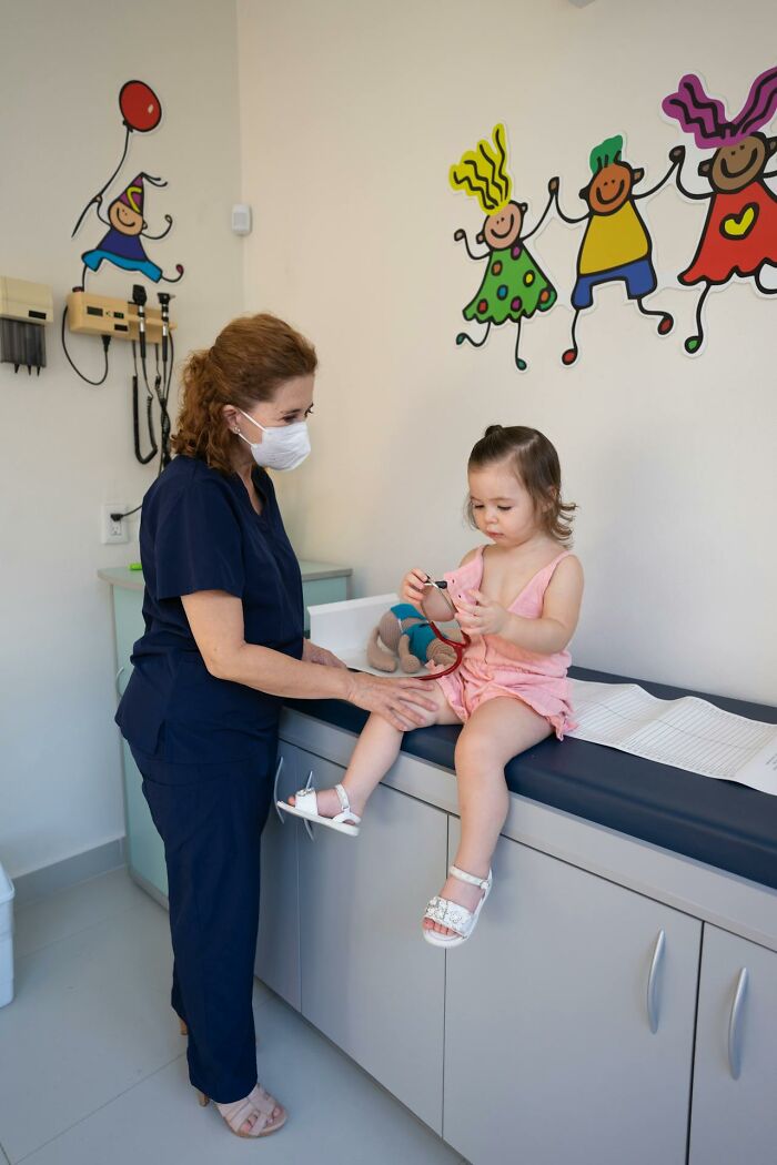 Pediatric nurse wearing mask attending to a young patient in a colorful medical exam room with playful wall art.