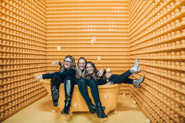 Three women sitting in a yellow bathtub surrounded by walls covered with yellow rubber ducks, promoting stay hydrated concept.