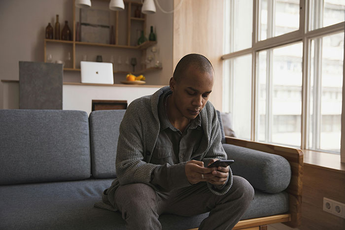 Man sitting on gray couch using phone indoors, depicting tension related to fiancé’s female friend demanding speech and best man role. - 8