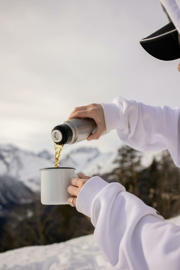 Person in a white sweatshirt pouring liquid from a thermos into a mug outdoors in a snowy mountain survival setting.