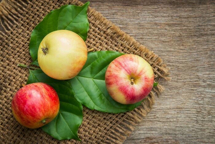 Three apples resting on large green leaves over a textured brown mat and wooden surface with natural lighting.