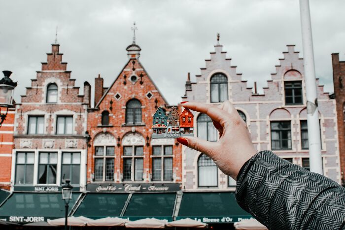 Hand holding miniature Brugge houses in front of traditional Belgian buildings, illustrating charming pickup lines setting.