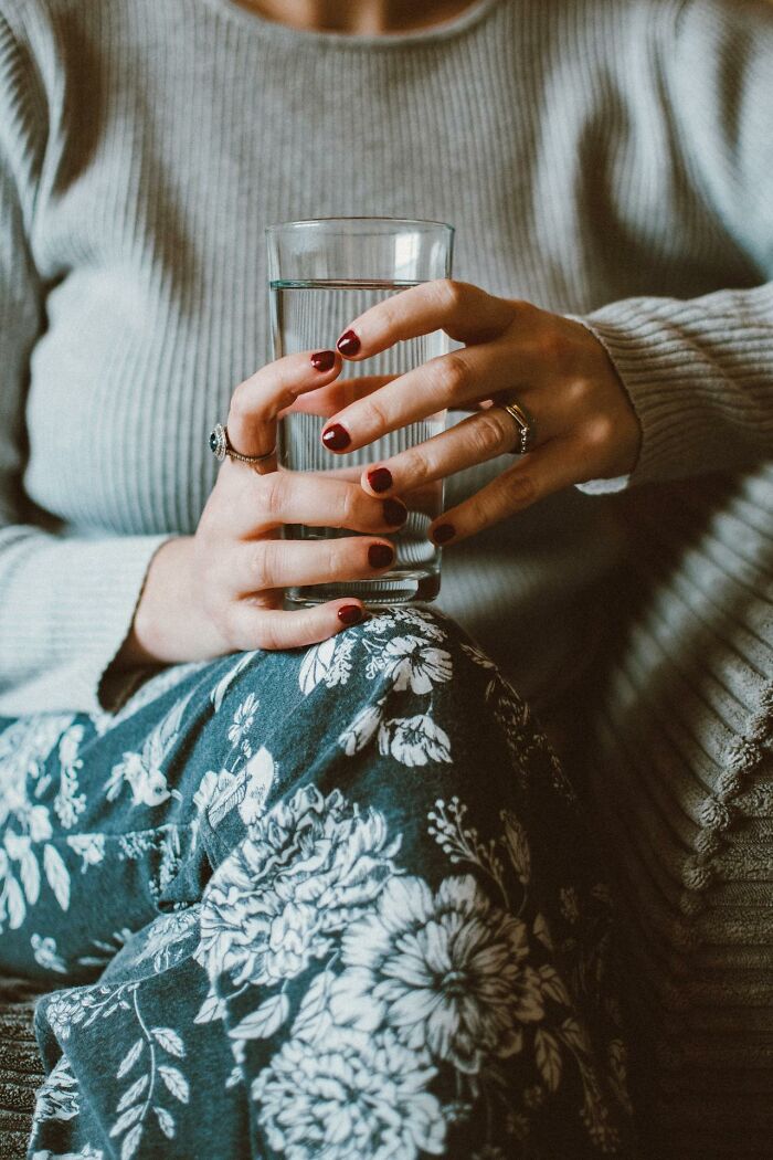 Person with red nail polish holding a glass of water while wearing a beige sweater and floral pants, promoting stay hydrated.