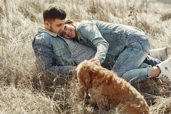 A young couple lying in dry grass, smiling and cuddling while a small dog plays nearby, showing relationship signs.