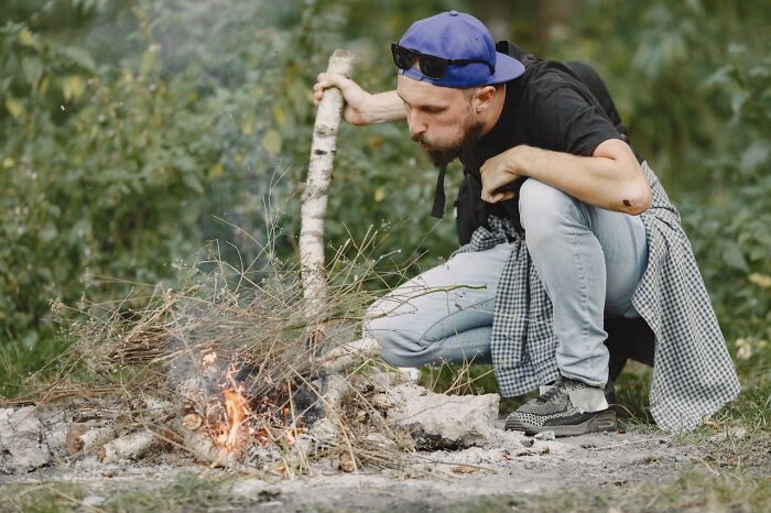 Man in a blue cap blowing on a small campfire in a forest setting illustrating survival tips concept.