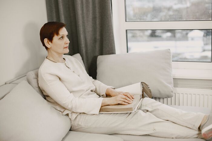 Middle-aged woman sitting on a couch reading a book, reflecting on harmless lies parents told that left her low key traumatized. - 9
