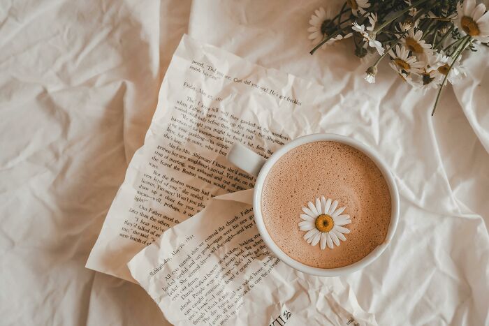 Cup of coffee with a daisy on froth resting on crumpled book pages and white fabric, evoking lost in life reflection.