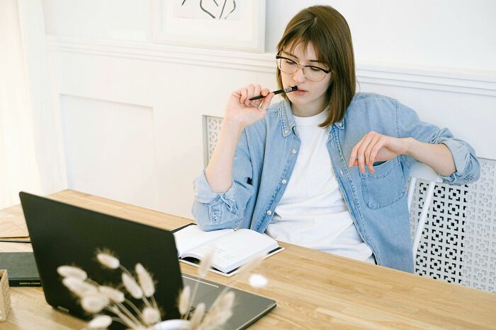 Young woman wearing glasses, thinking while solving 39 true-or-false questions on a laptop at a wooden desk.