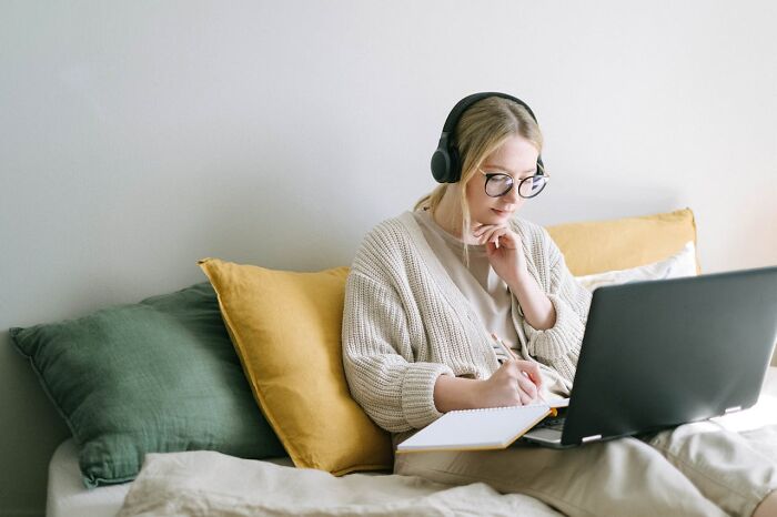 Woman wearing headphones taking notes while using a laptop on a bed with green and yellow pillows, stay hydrated concept.