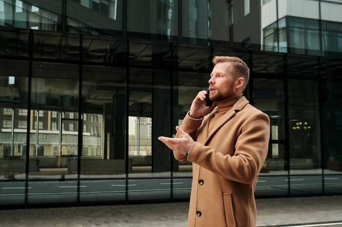 Man in a tan coat speaking on phone outside modern glass building, capturing disturbing things people overheard conversation.