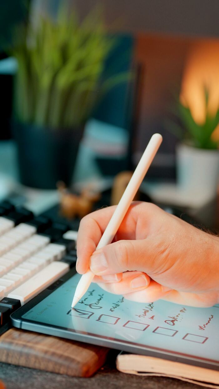 Hand holding stylus writing a checklist on a tablet, illustrating stay hydrated reminders in a cozy workspace.
