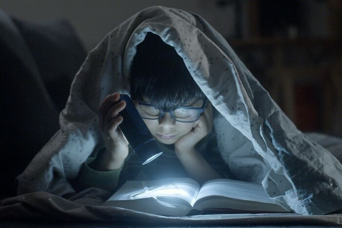 Child reading a book under a blanket using a flashlight, illustrating harmless lies from parents that left trauma. - 25