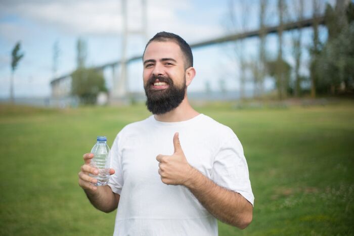 Bearded man outdoors holding a water bottle and giving a thumbs up, promoting stay hydrated for better health.