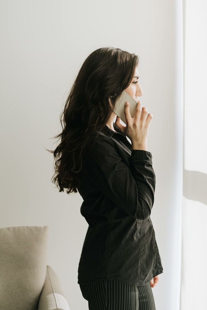 Woman with long dark hair wearing black shirt talking on phone by window, illustrating disturbing things overheard conversation.