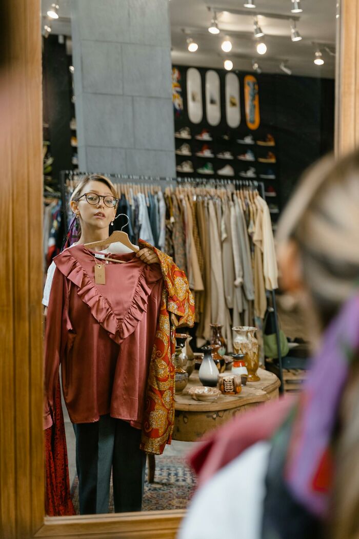 Woman holding a blouse in front of a mirror, reflecting on toxic best friends and personal choices in a clothing store. - 10