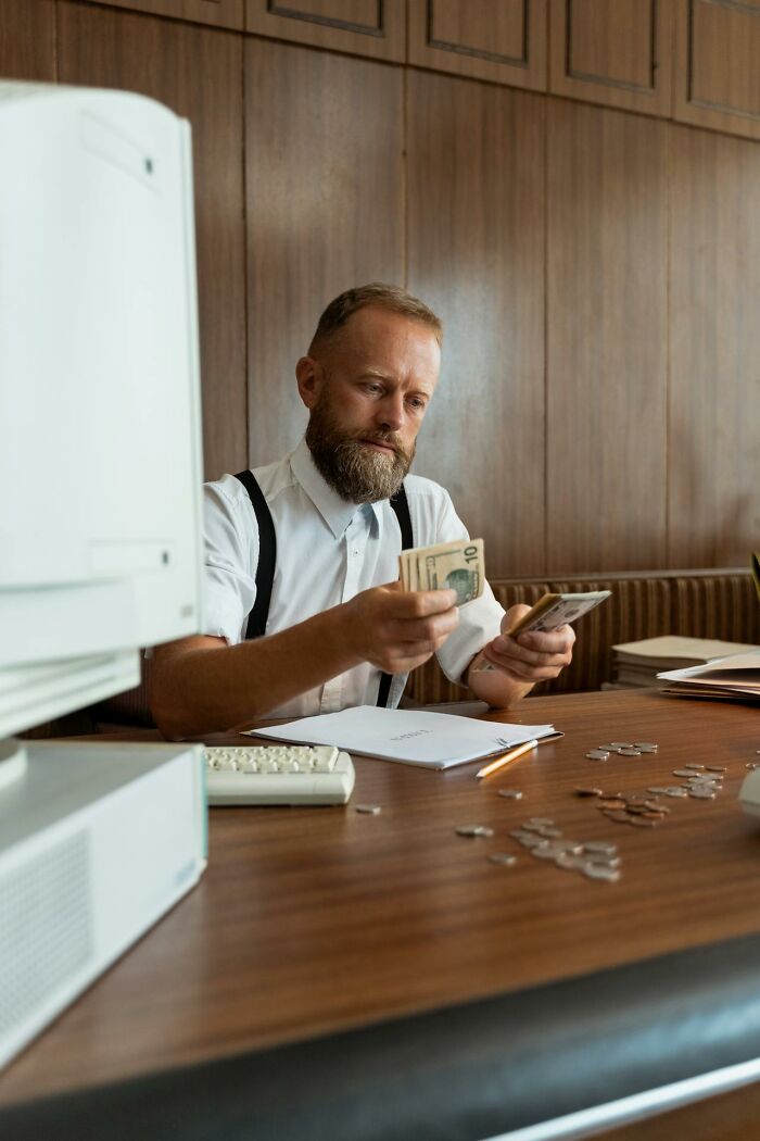 Older man counting money at desk, reflecting on life after divorce in a vintage office setting with wooden panels. - 25