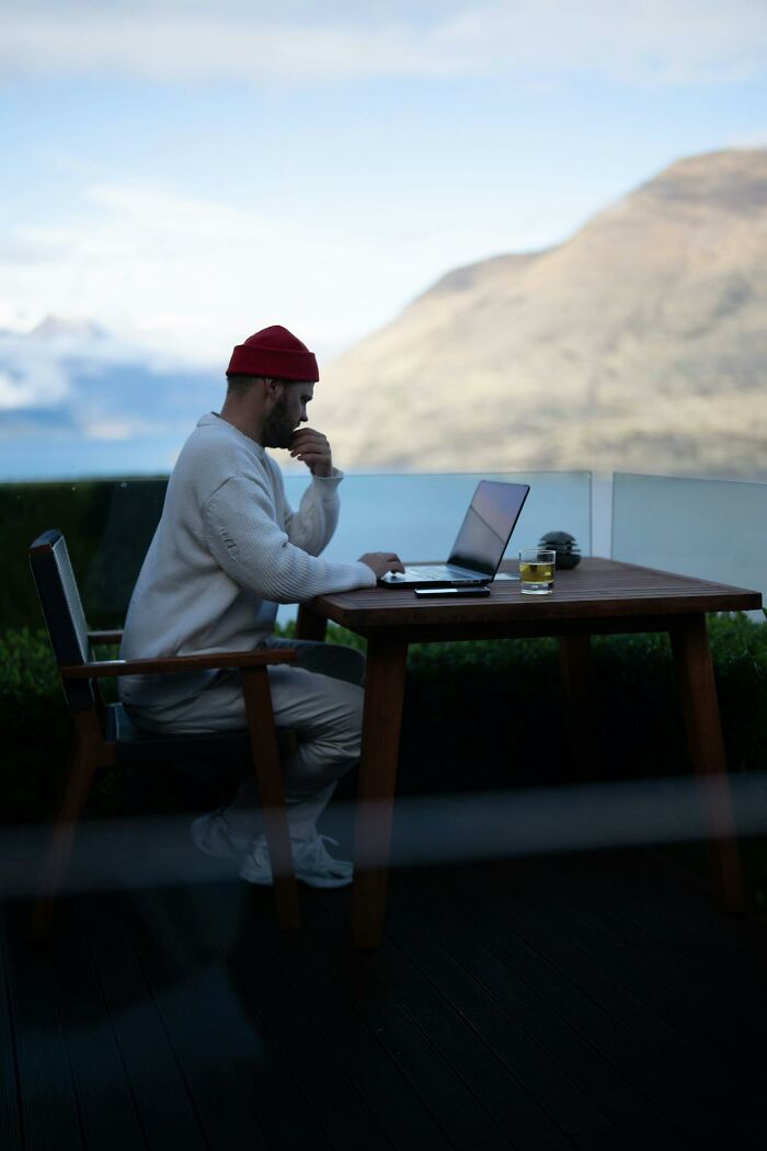 Man wearing a red beanie working on a laptop outdoors with a glass of water to stay hydrated on a wooden table.