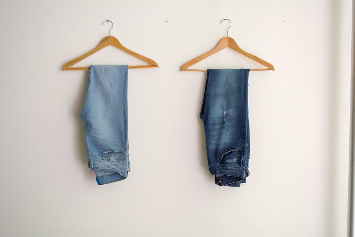 Two pairs of folded jeans hanging on wooden hangers against a plain white wall in a minimal setting.