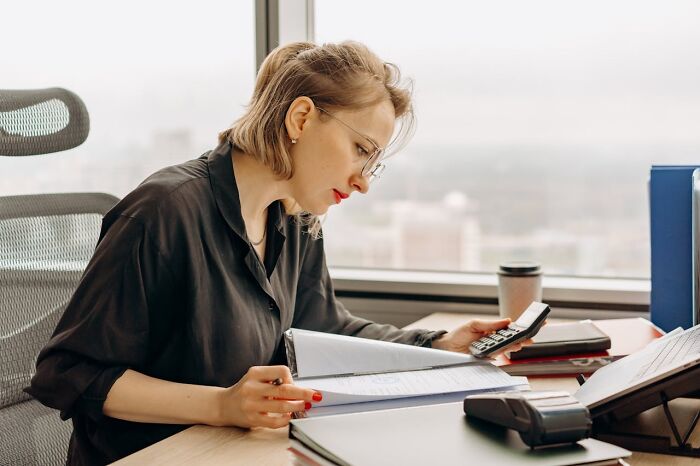 Woman working at office desk, using calculator and reviewing documents, focused on jobs that make 6 figures income.