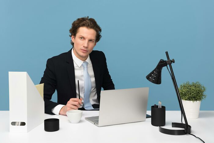 Young man in a suit at desk with laptop and lamp, playing guess who with Wikipedia descriptions of notable people.