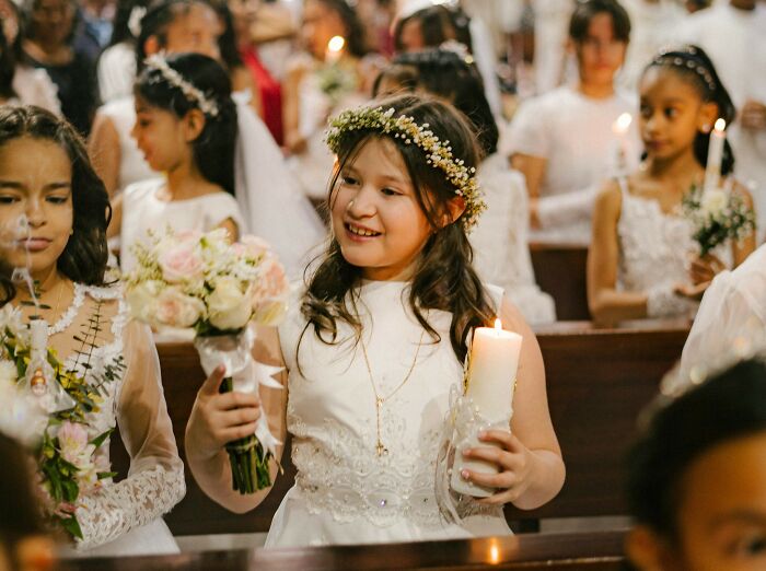 Young students dressed in white dresses holding flowers and candles during a ceremonial event in a church setting.