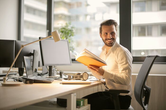 Man smiling while reading a book at his desk in a modern office, with red flag behaviors subtly highlighted. - 28
