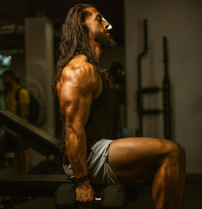 Muscular man with long hair working out with dumbbells, showing strength and focus in a dimly lit gym environment.