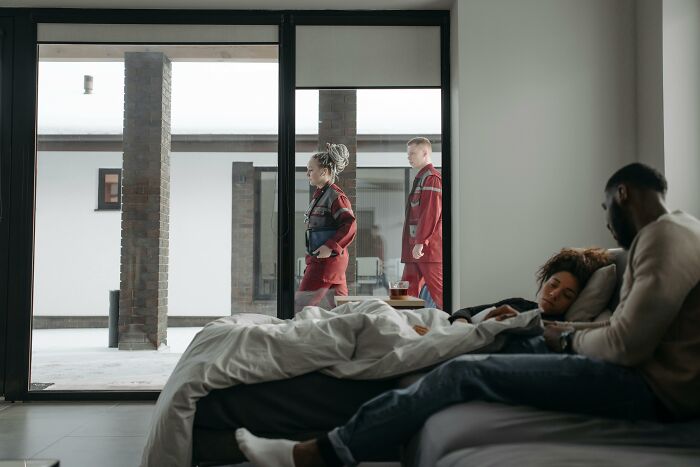 Two paramedics walk outside a modern home while a man sits beside a resting patient in the bedroom.