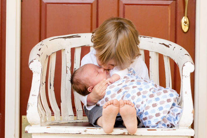 Young boy gently kissing baby wrapped in blanket while sitting on rustic white wooden bench in front of red door. - 12