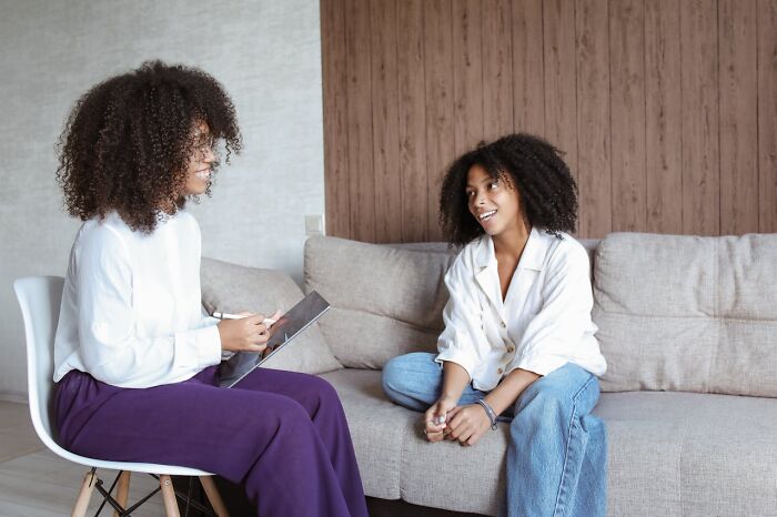Two women with curly hair talking on a couch and chair in a casual setting emphasizing the importance of staying hydrated.