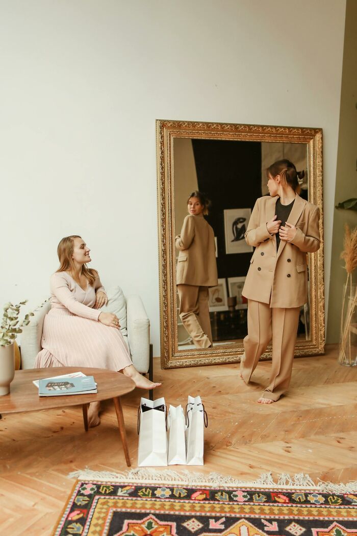 Two women in a cozy room, one sitting and one standing near a large mirror reflecting toxic best friends behavior. - 6