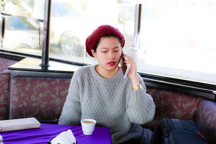 Young woman in a red beanie and gray sweater talking on phone at cafe discussing pettiest things breakups borderline insane