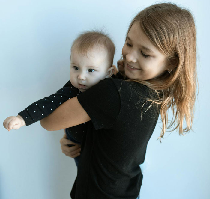 Teen babysitter holding a baby, smiling softly against a plain background, reflecting babysitting and teen topics.