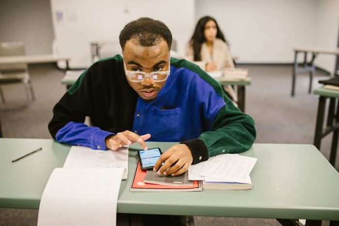 Student cheating on phone during a school exam, surrounded by papers and books in a classroom setting. - 8