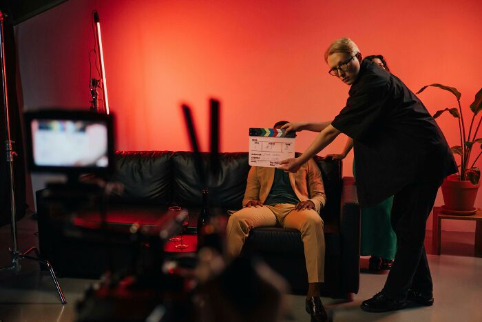 Film crew member holding clapperboard in front of actor on sofa, capturing main character energy on set with red lighting. Film crew member holding clapperboard in front of actor on sofa, capturing main character energy on set with red lighting.