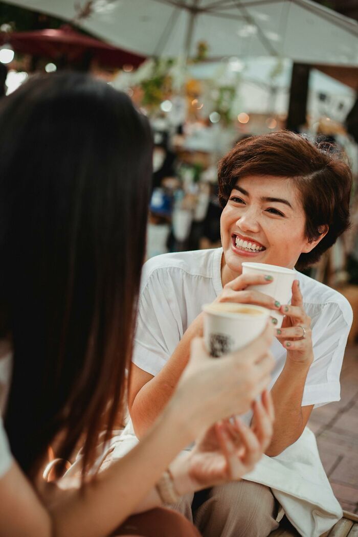 Two women smiling and holding coffee cups outdoors, engaged in a casual conversation with disturbing things overheard.