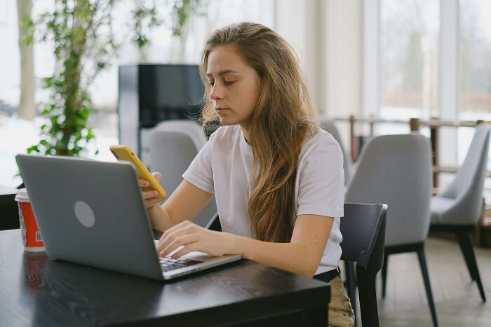 Young woman focused on her phone and laptop in a cafe, illustrating pettiest things causing breakups borderline insane scenarios.