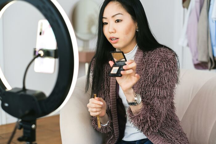 Young woman applying makeup on camera with ring light, illustrating trends leading to overconsumption in beauty content creation. - 3