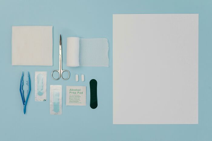 First aid kit items including scissors, gauze, alcohol prep pad, pills, and tweezers neatly arranged on blue background.