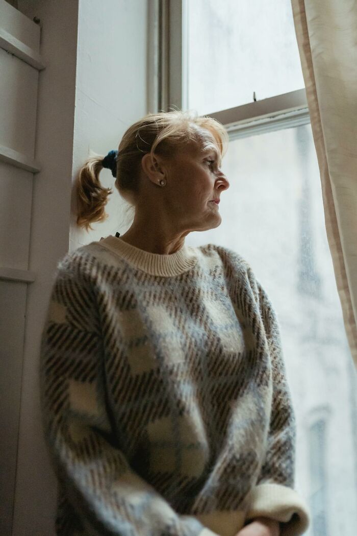 Older woman looking out window, reflecting on life after divorce, wearing cozy patterned sweater indoors. - 32