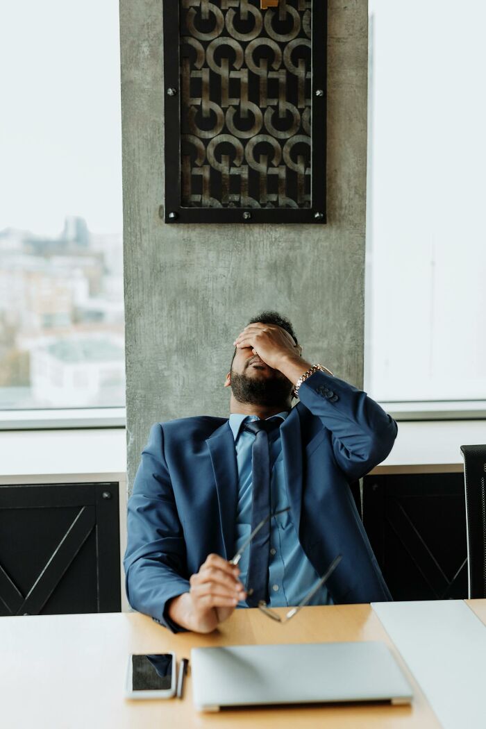 Stressed man in blue suit at office desk, holding glasses and covering face, reflecting workplace revenge and frustration. - 15