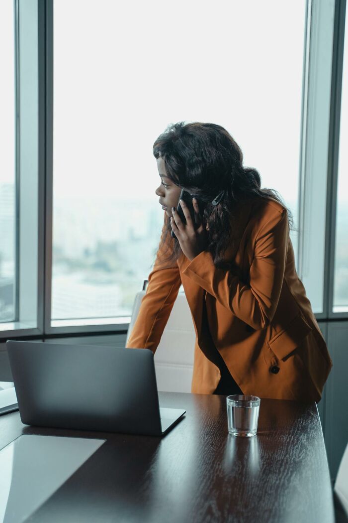 Woman in orange blazer talking on phone, standing by laptop in office with large windows, showing cheating red flags concept. - 14