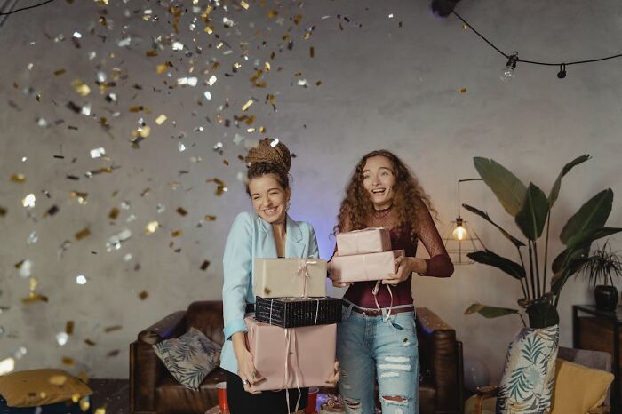 Two young women smiling and holding gift boxes indoors with confetti falling, illustrating toxic best friends awareness. - 52