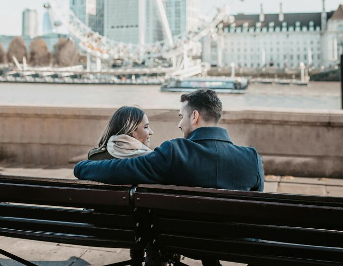 Couple sitting on a bench by the river with London Eye in the background, discussing truly outrageous student names.