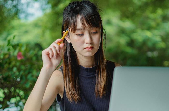 Young woman looking thoughtful, holding a pencil near her face while working on a laptop in a natural setting. - 65