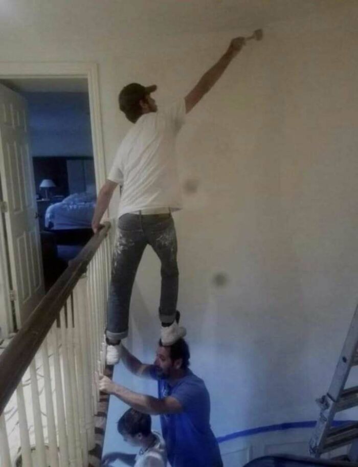 Man standing on another person's shoulders painting a wall, illustrating hilariously dumb engineering mistakes at home.