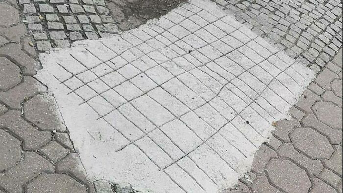 Uneven pavement repair with chaotic concrete patch blending trash and culture in a humorous urban setting.