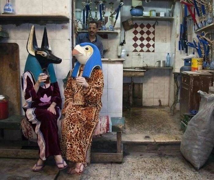 Two people wearing animal head masks sit in a cluttered room, blending trash and culture in a chaotic scene.