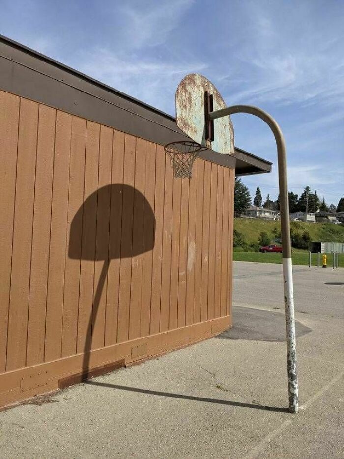 Rusty basketball hoop bent awkwardly with shadow showing a proper hoop shape, a funny engineering mistake outdoors.